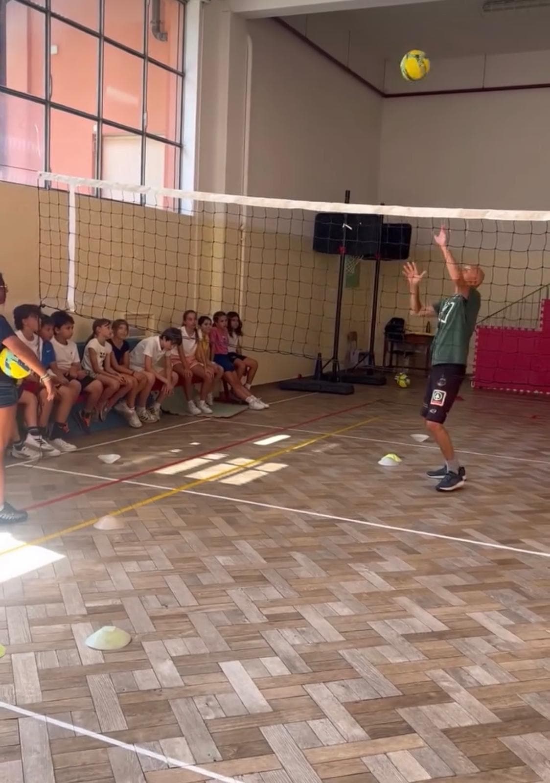 Uomo che gioca a pallavolo in una palestra davanti a bambini seduti in panchina.
